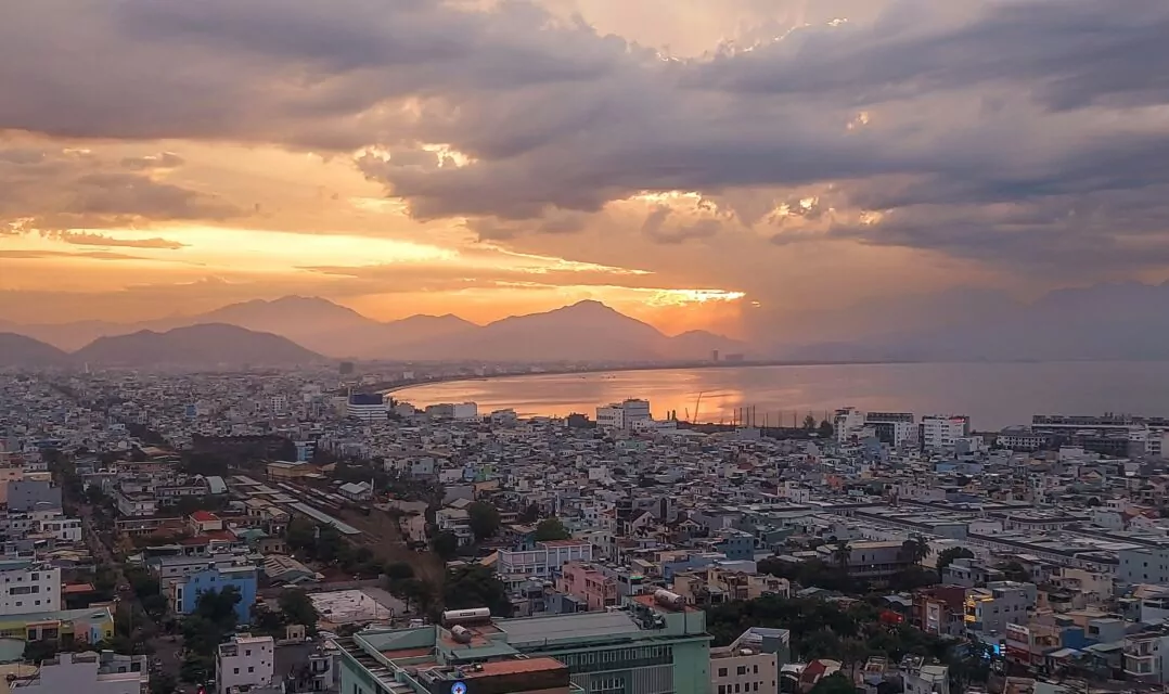 View over Đà Nẵng towards the evening sun setting in warm colors behind the hills and reflecting in the water of Đà Nẵng Bay. Đà Nẵng has a population of about 1.134 million and is located just under 800 kilometers north of Sài Gòn on the coast.