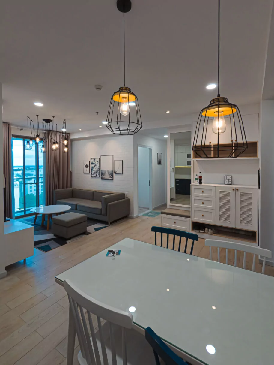 Modern apartment with a cozy living and dining space, featuring wooden floors, a grey sofa, glass-top table, and pendant lighting. Large windows offer a city view, and minimalist decor adds a stylish touch.
